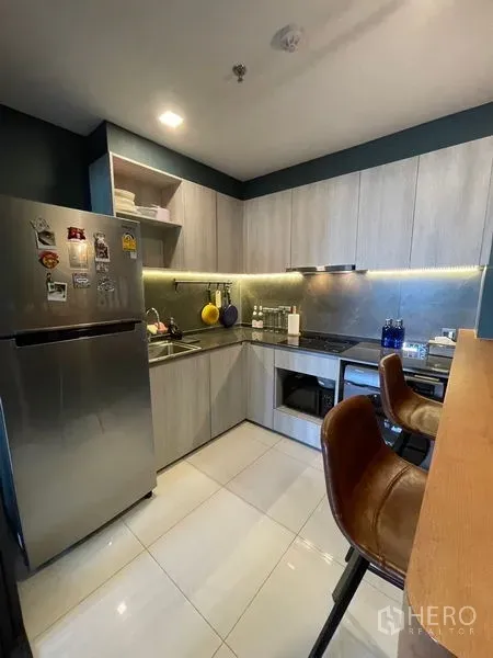 คอนโด ขาย - ครัวรูปตัว L โทนไม้ มีตู้บิวท์อิน ไฟใต้ตู้ เคาน์เตอร์บาร์ และตู้เย็นสแตนเลส