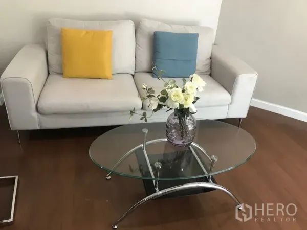 Condominium for rent or sale in Huai Khwang, Bangkok - Living room with light sofa, colorful cushions and oval glass coffee table with flowers.