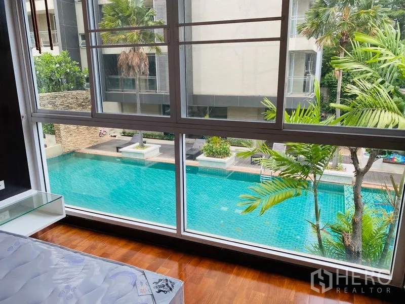 Condominium for rent in Watthana, Bangkok - Large window view of a turquoise swimming pool and palm trees in the courtyard.
