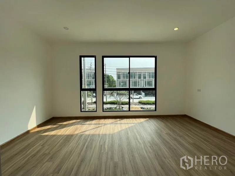 Townhouse for rent in Bang Kapi, Bangkok - Sunlit bedroom with tall windows and street-facing view of neighboring townhouses.