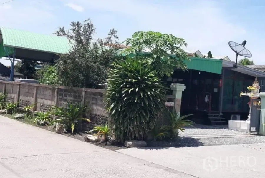Land for sale in Mueang Samut Prakan, Samut Prakan - Walled land plot with landscaped plants, a gravel driveway and green-roofed outbuildings along a quiet concrete road.
