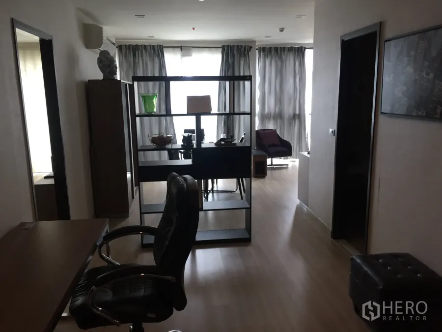 Condominium for rent in Watthana, Bangkok - Open-plan living area with light wood floors, shelving divider, work desk, and large windows with sheer curtains in a Bangkok condo.