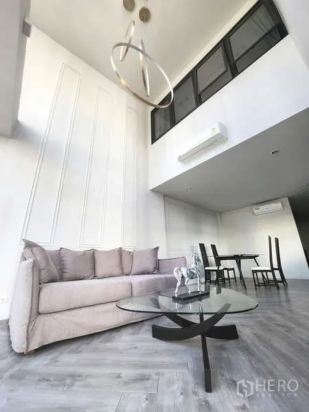 联排别墅 出租在Watthana, Bangkok - 挑高客厅，灰色沙发与玻璃茶几，现代吊灯，连接用餐区。