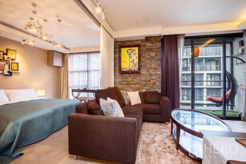 公寓 出租在Watthana, Bangkok - 客厅通往阳台，转角沙发与玻璃桌，旁为阁楼风卧室与石纹装饰墙。