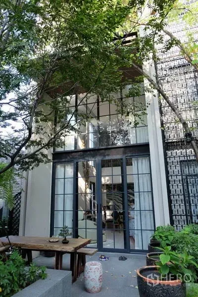 Townhouse for rent in Khlong Toei, Bangkok - Garden patio with trees facing a double‑height townhouse facade and black‑framed glass doors.