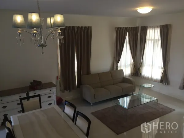 联排别墅 出租在Watthana, Bangkok - 明亮客厅，米色沙发、玻璃茶几与带窗帘的转角窗。