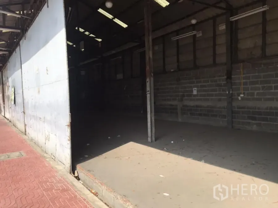 Warehouse for rent in Don Mueang, Bangkok - Open-sided warehouse with high roof, concrete floor, brick walls and fluorescent lighting beside a red-paved sidewalk in Don Mueang.