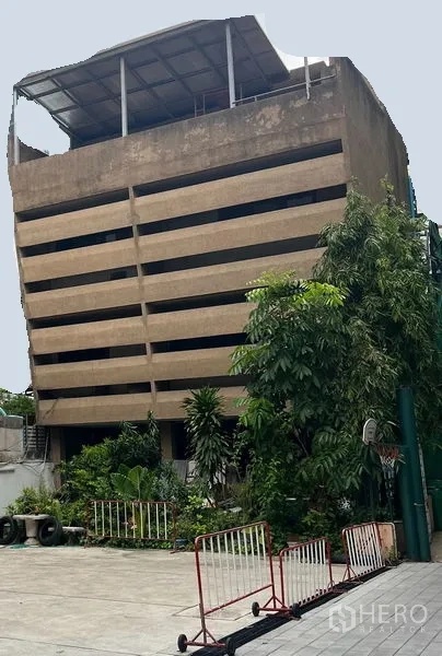 พื้นที่ขายปลีก ให้เช่าในKhlong Toei, Bangkok - อาคารคอนกรีตสูงหลายชั้น หน้าระแนงแนวนอน มีหลังคาดาดฟ้า และลานกว้างด้านหน้าพร้อมต้นไม้และแผงกั้น ในเขตคลองเตย กรุงเทพฯ