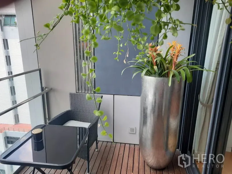 Condominium for rent or sale in Sathon, Bangkok - Private balcony with wood decking, glass railing, a small table and a tall planter with tropical greenery.