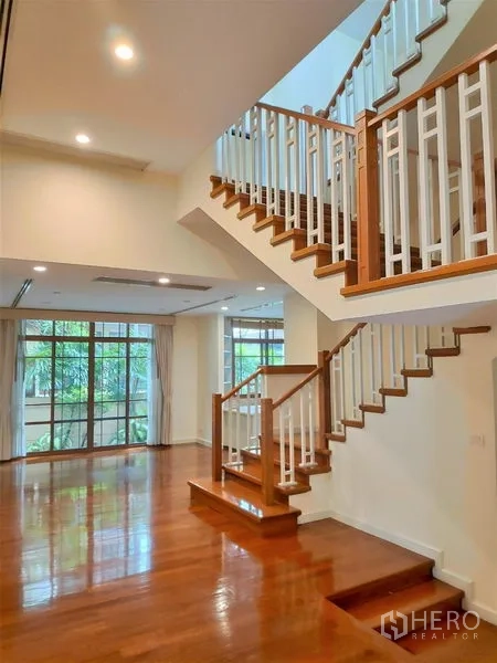 独立式住宅 出租在Watthana, Bangkok - 通透的双层挑空空间，木质楼梯与大窗，面向花园景观。