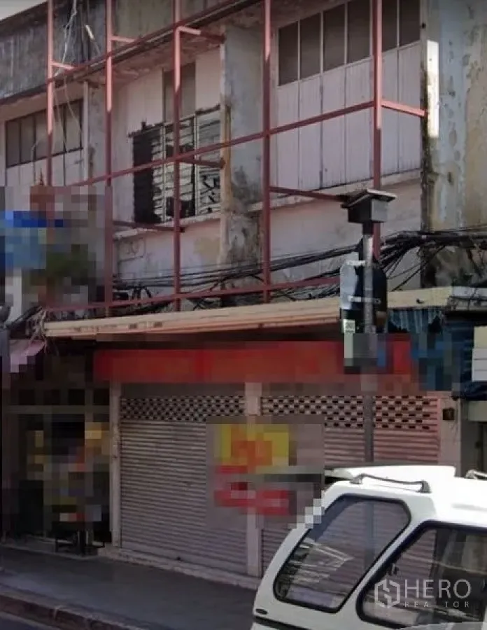Shophouse for rent in Sathon, Bangkok - Street view of an older multi-storey shophouse in Sathon, Bangkok, with a closed metal roller shutter, weathered facade, red steel sign frame and overhead cables.