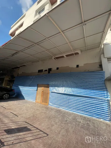 零售空间 出租在Watthana, Bangkok - 瓦他那区 M Towers 一层临街商铺外立面，宽大雨棚、蓝白条纹临时围挡、木质双开门与前方停车区。