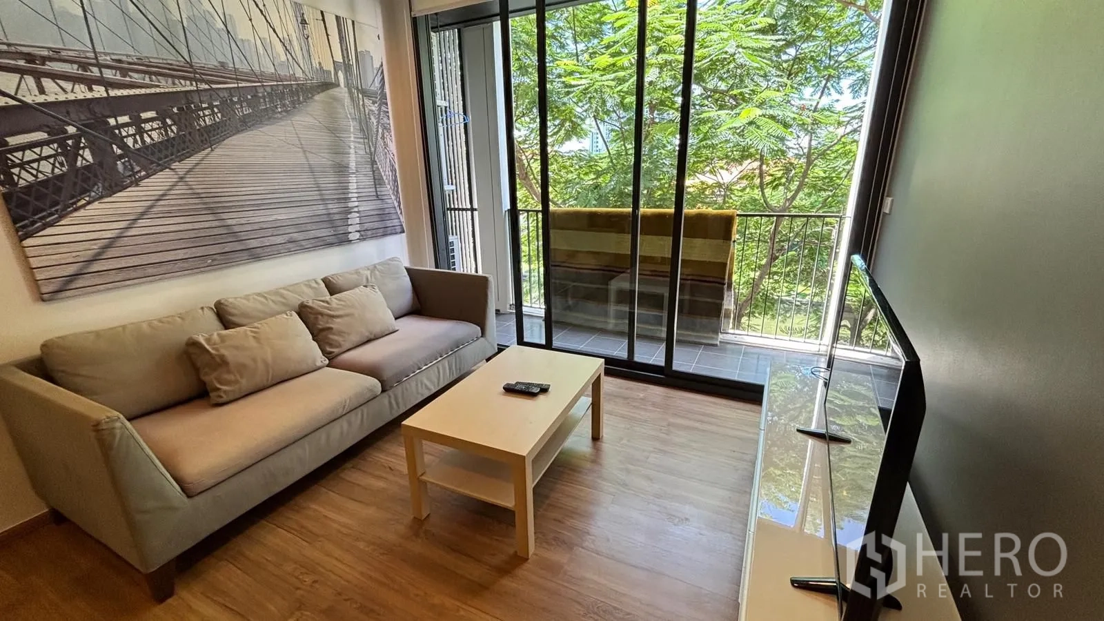 Condominium for rent or sale in Watthana, Bangkok - Bright living space with large artwork, coffee table, and full-height windows to a green view.