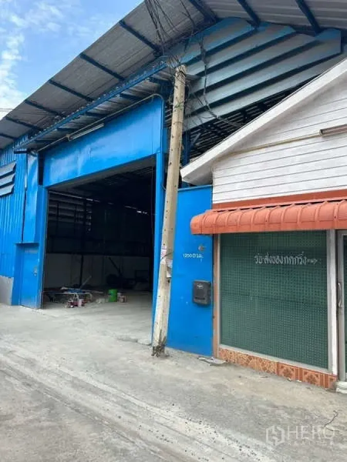货栈 出租在Prawet, Bangkok - 曼谷帕拉威区蓝色金属仓库外观，宽幅卷闸门开启，旁侧为小型办公室，临窄混凝土巷道。