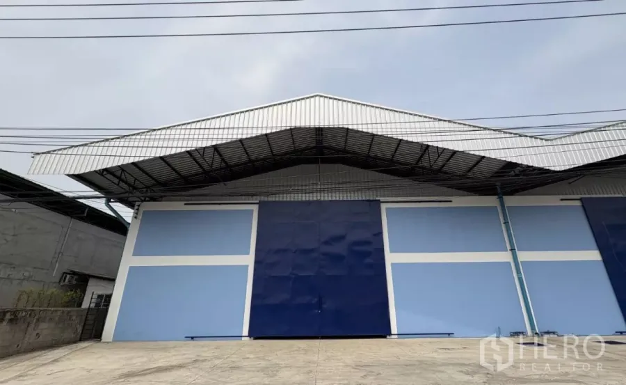 Warehouse for rent in Mueang Samut Prakan, Samut Prakan - Street-facing facade of a new warehouse with wide blue sliding doors and a metal awning roof.