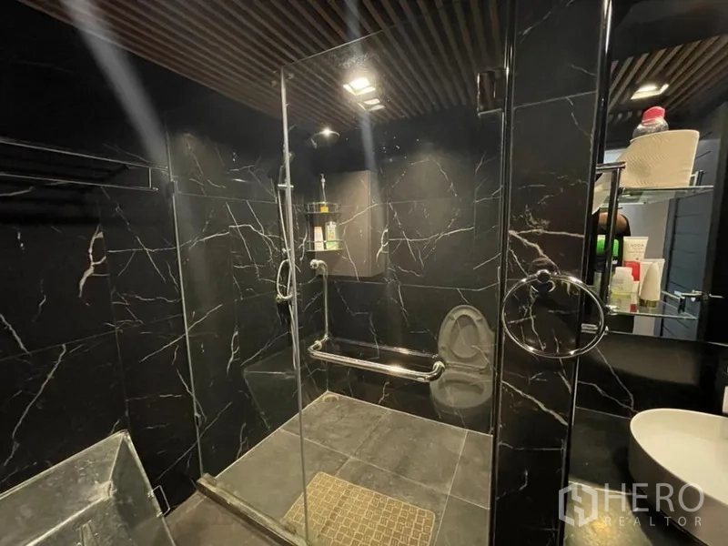 Condominium for sale in Chatuchak, Bangkok - Glass-enclosed shower with rain head and black marble walls in a modern bathroom.