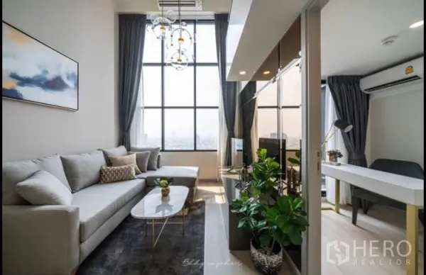 Condominium for rent in Sathon, Bangkok - Bright Sathon condo living room with double-height windows, gray sectional sofa, TV wall and a work desk.