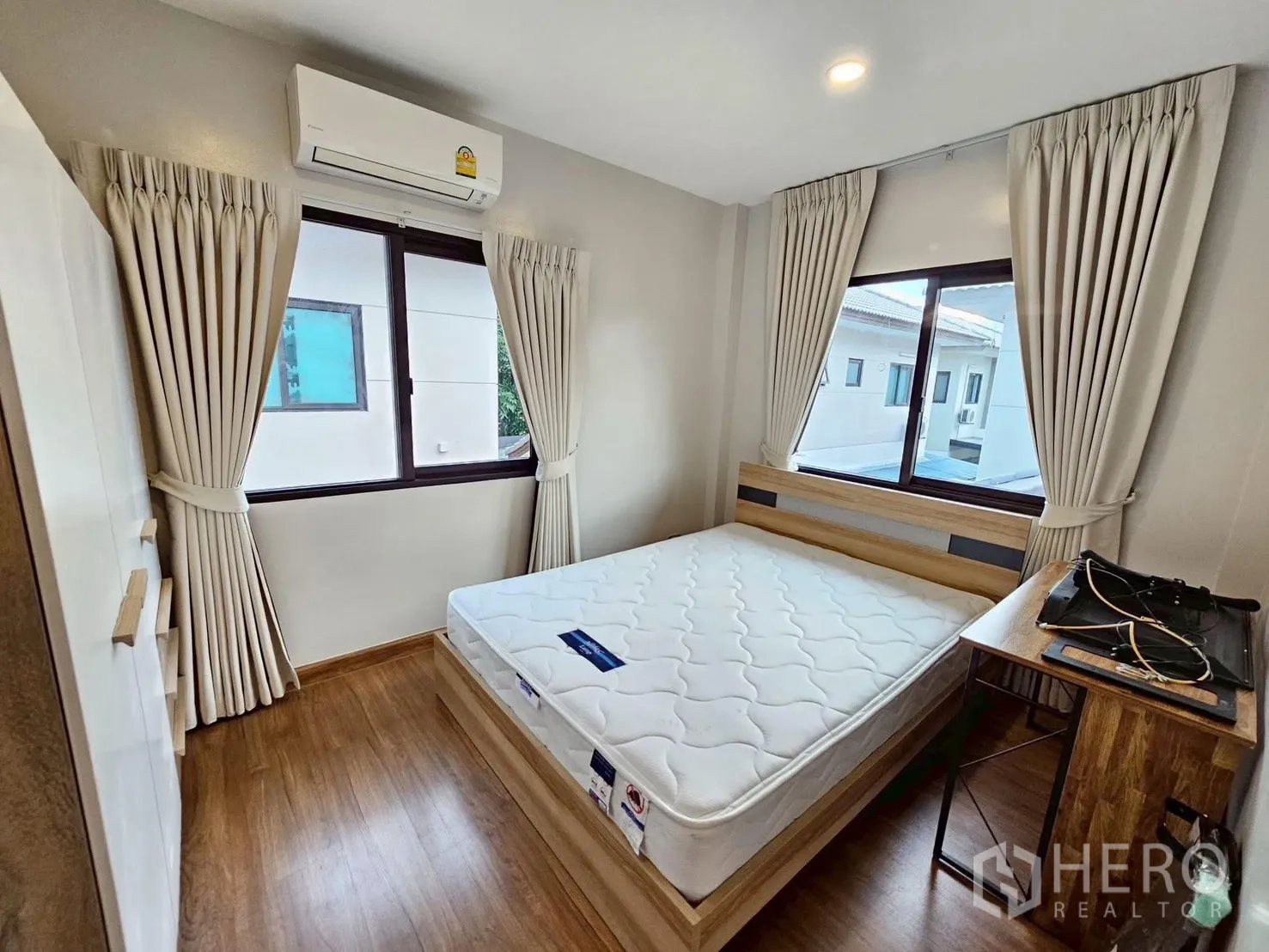 Detached House for sale in Bang Phli, Samut Prakan - Compact bedroom with double bed, study desk, two large windows with curtains, wood floors, and air conditioning.