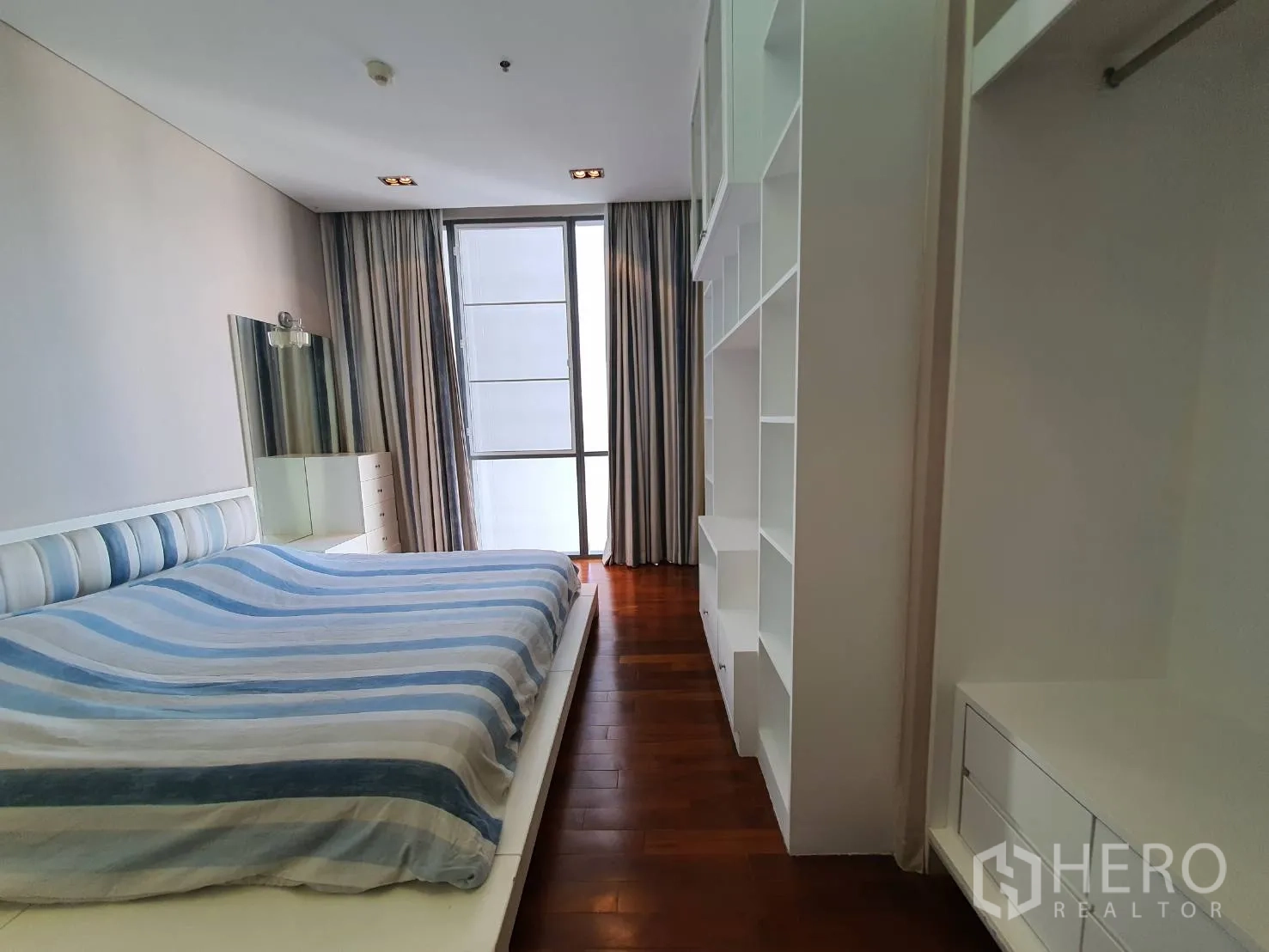 Condominium for rent in Khlong Toei, Bangkok - Bedroom with striped bedding, open shelving and a floor-to-ceiling window.
