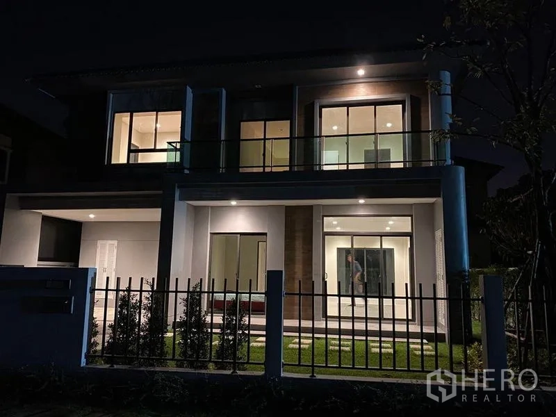 Detached House for rent - House exterior at night with warm lighting, balcony and fenced garden.