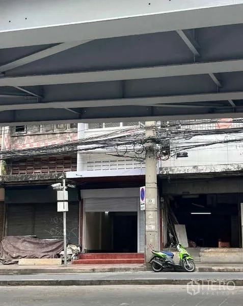 店屋 出租在Din Daeng, Bangkok - 丁登区临街商铺外立面，卷闸门入口与红色台阶，上方电线密集，立交桥下停有一辆摩托车。