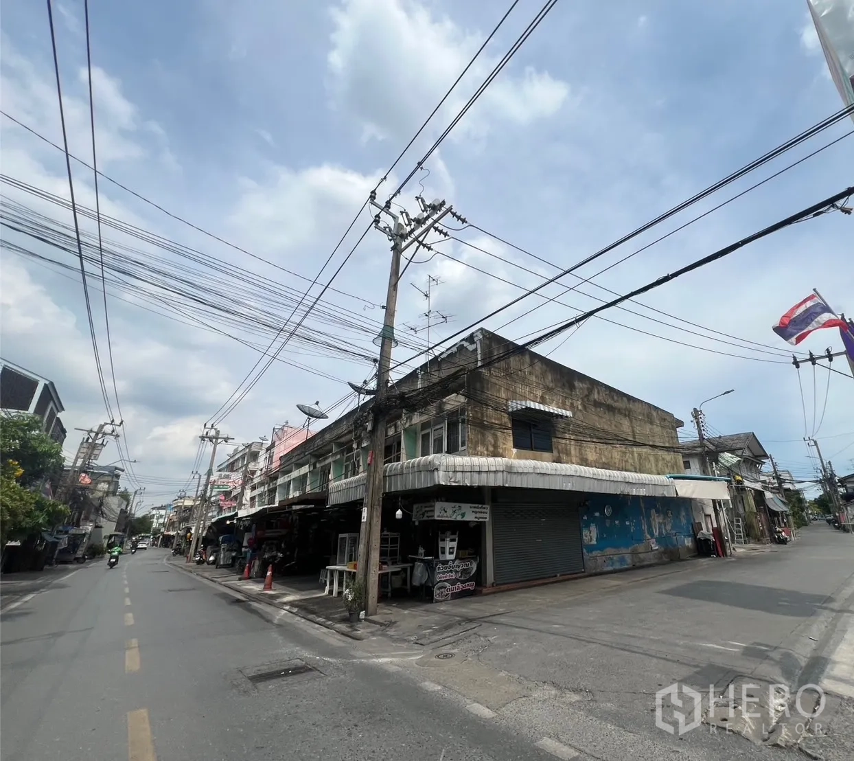 ที่ดิน ให้เช่าหรือขายในPhra Khanong, Bangkok - แยกหัวมุมอาคารพาณิชย์สองชั้นบนถนนย่านพระโขนง มีสายไฟเหนือศีรษะและธงชาติท่ามกลางท้องฟ้าโปร่ง.