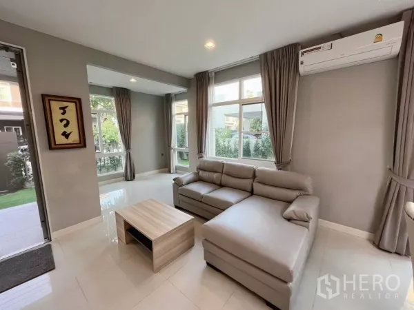 Detached House for rent in Samut Prakan - Bright living room with L-shaped sofa, large windows, and glossy tiled flooring.