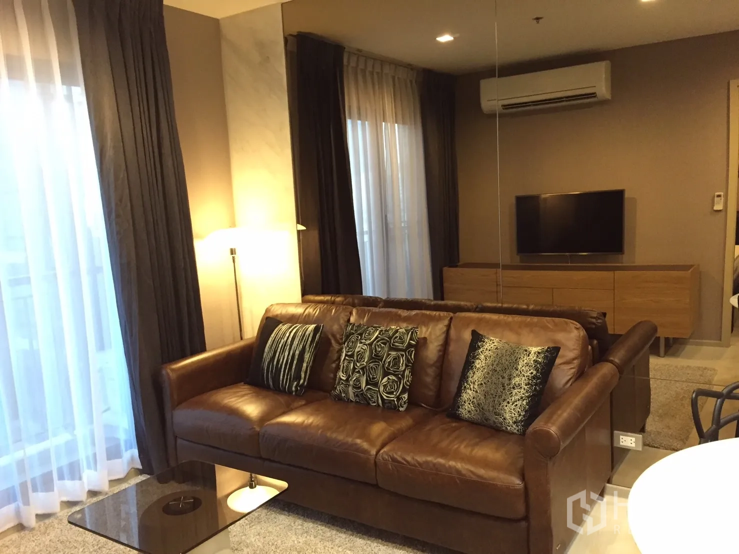 คอนโด ให้เช่าในKhlong Toei, Bangkok - ห้องนั่งเล่นโทนอุ่น โซฟาหนังสีน้ำตาล โคมไฟ ชั้นวางทีวี และหน้าต่างผ้าม่านบานใหญ่