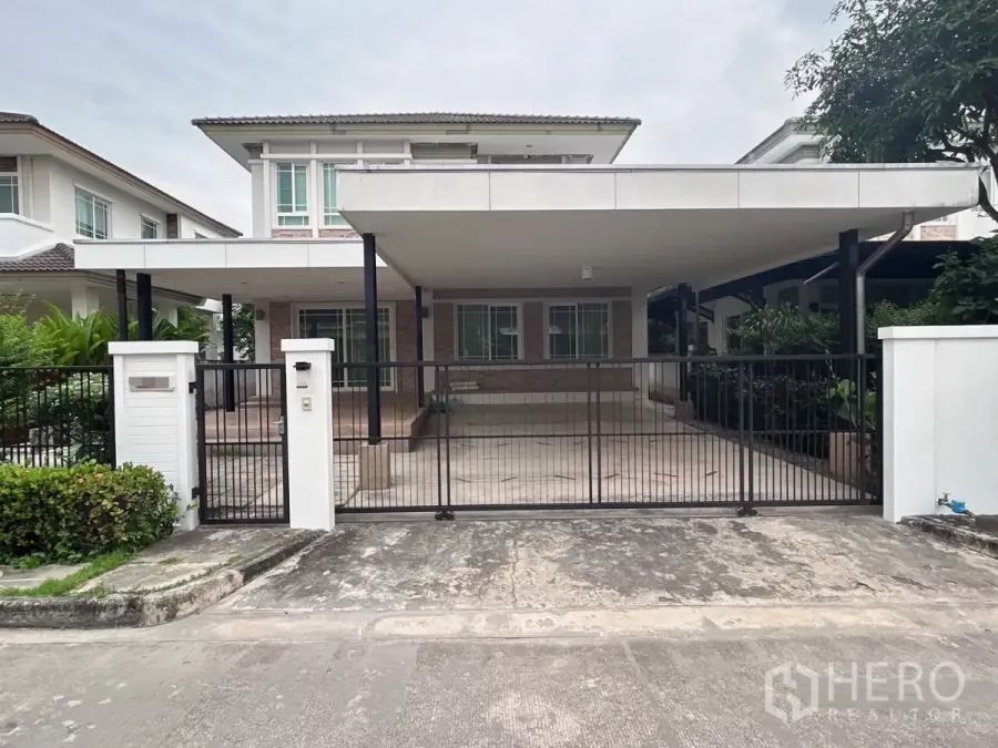 บ้าน ขายในBang Kapi, Bangkok - มุมหน้าบ้านเดี่ยวสองชั้นพร้อมหลังคาจอดรถกว้างและประตูรั้วเหล็กในหมู่บ้านจัดสรร