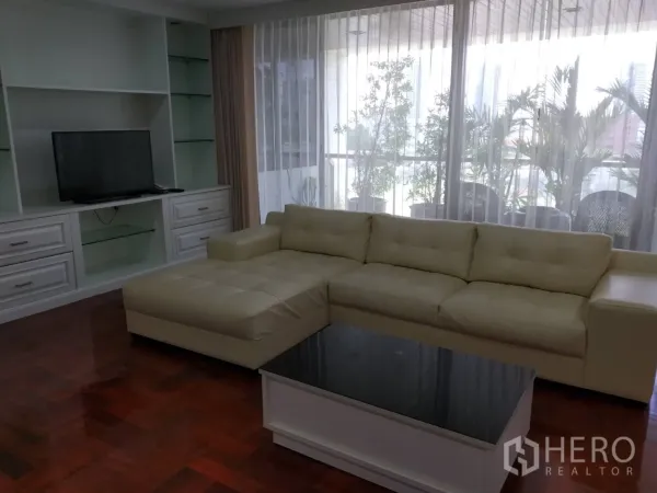 Condominium for rent in Pathum Wan, Bangkok - Living area with cream L-shaped sofa, built-in TV cabinet and balcony greenery behind sheer drapes.