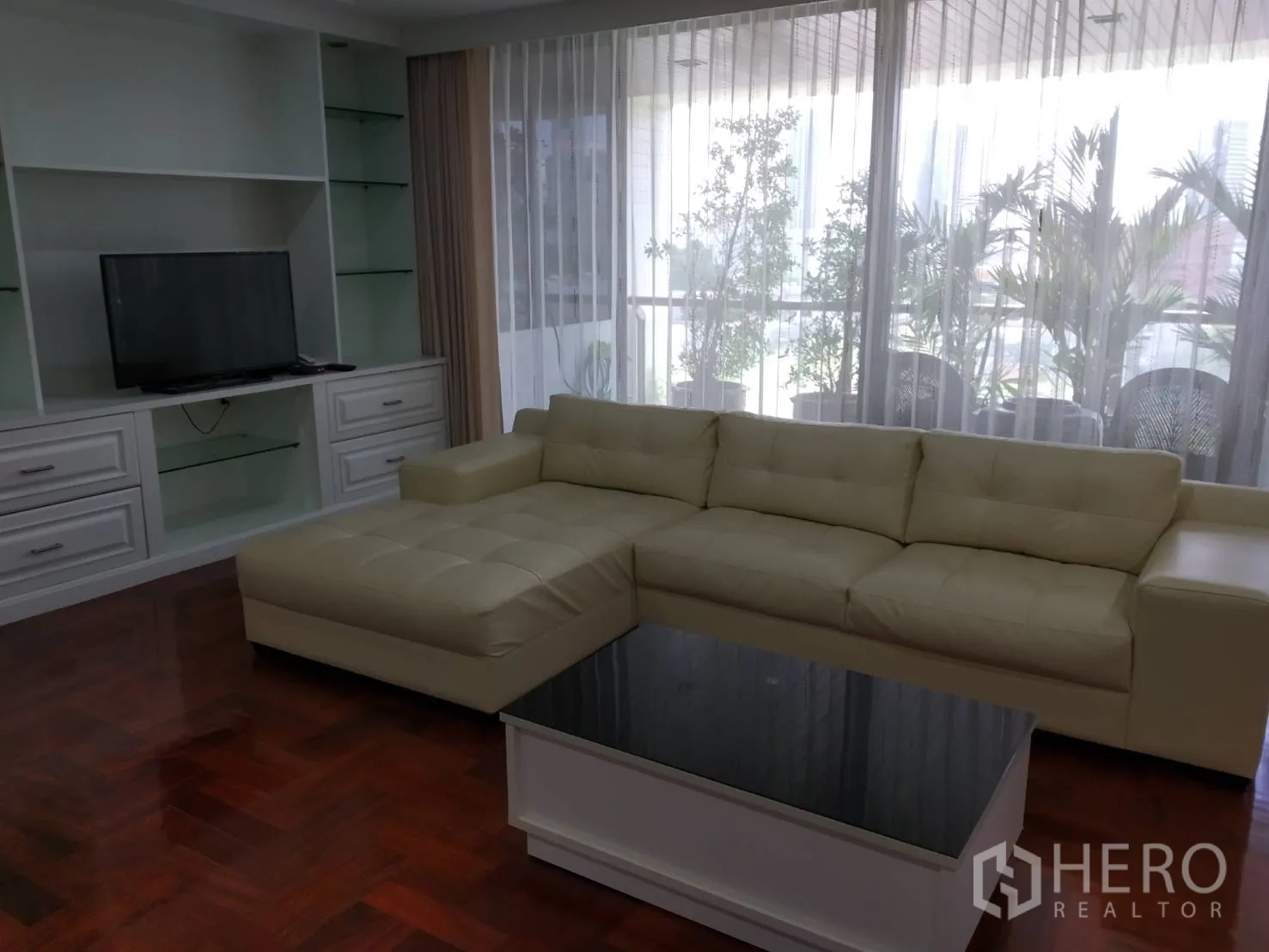 Condominium for rent in Pathum Wan, Bangkok - Living area with cream L-shaped sofa, built-in TV cabinet and balcony greenery behind sheer drapes.