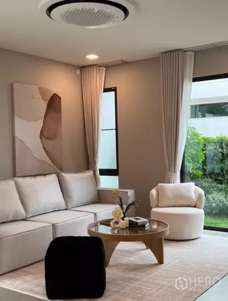 Detached House for rent in Samut Prakan - Cozy living room with L-shaped sofa, round coffee table, tall windows, curtains and lush garden backdrop.