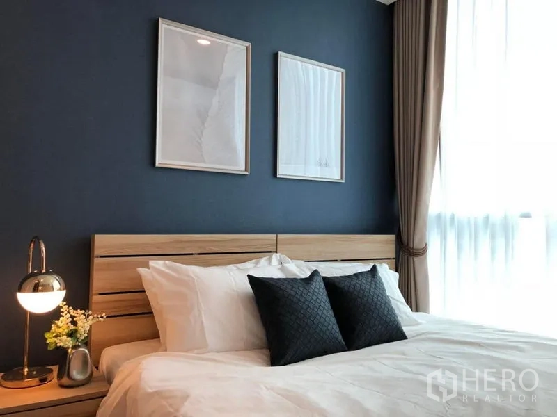 คอนโด ขาย - ห้องนอนหลักสว่าง ผนังสีน้ำเงิน เตียงไม้ ผ้าปูสีขาว และหน้าต่างเต็มบานพร้อมม่านโปร่ง
