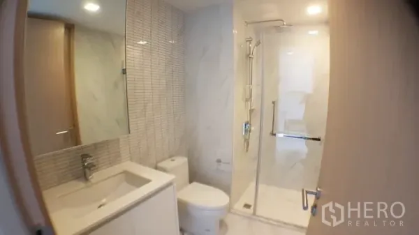 Condominium for rent in Watthana, Bangkok - Modern bathroom in the condominium with glass-enclosed shower, large mirror and white marble tiles.