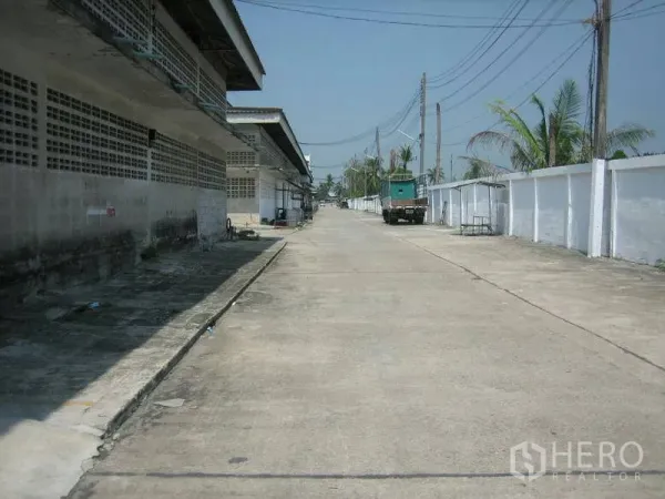 货栈 出租在Min Buri, Bangkok - 工业园区内宽阔混凝土通道，两侧为仓库建筑，电线与停放的卡车可见