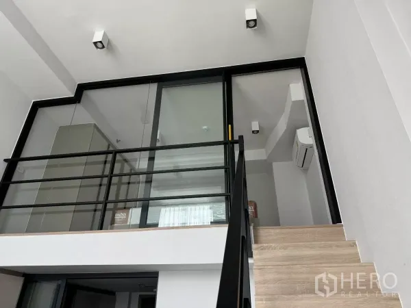 Condominium for rent in Huai Khwang, Bangkok - Loft mezzanine with glass partition and black metal railing accessed by wooden stairs.