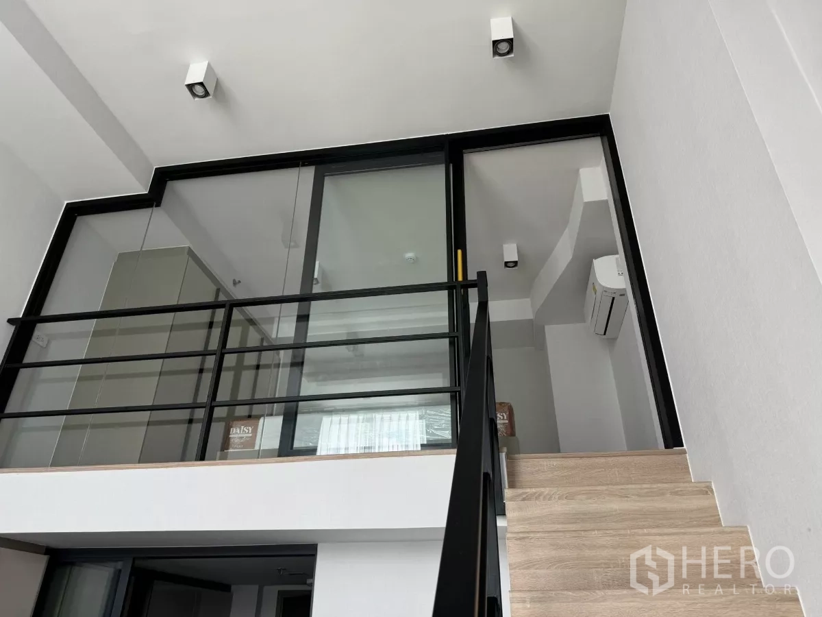Condominium for rent in Huai Khwang, Bangkok - Loft mezzanine with glass partition and black metal railing accessed by wooden stairs.