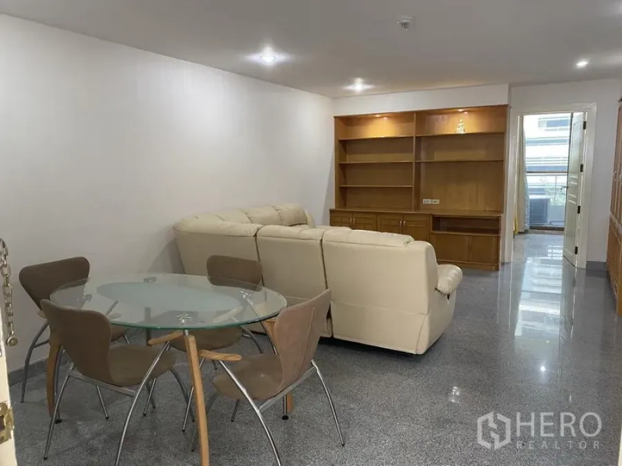 Condominium for rent or sale in Watthana, Bangkok - Dining and lounge area with a round glass table, four chairs, cream sofa and a built-in TV wall unit under recessed lights.