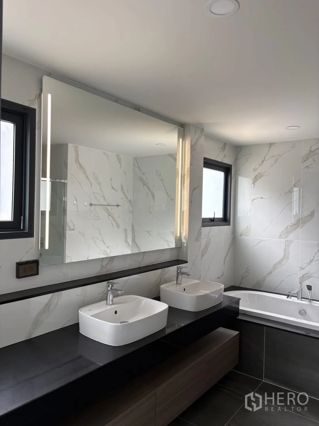 Townhouse for rent - Close view of double sink vanity with lighted mirror and bathtub on a stone counter.