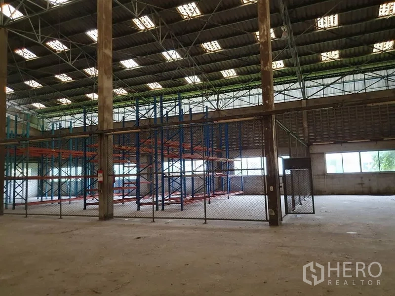 Factory for rent in Samut Prakan - Warehouse bay featuring blue and orange pallet racking behind chain-link fencing under bright skylight panels.