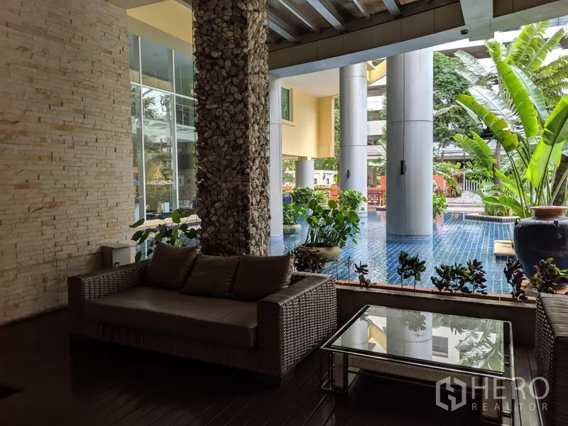 Condominium for rent in Watthana, Bangkok - Shaded outdoor lounge with rattan sofa, glass table and tropical plants beside the pool.