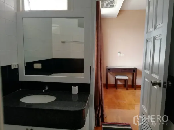 Condominium for rent in Watthana, Bangkok - Bathroom vanity with granite countertop and big mirror, looking out to a room with desk and curtains.