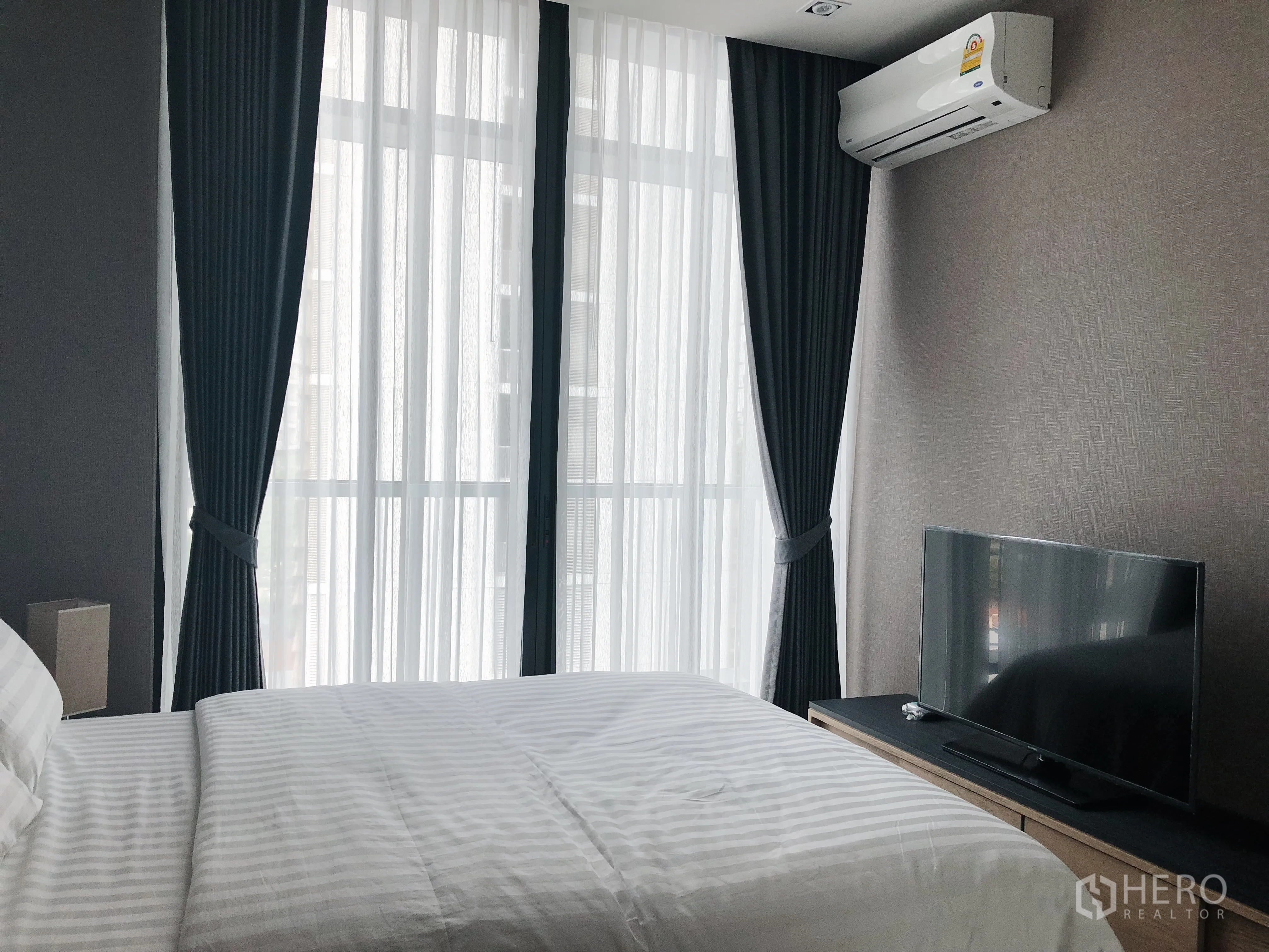 Condominium for rent in Khlong Toei, Bangkok - Bright bedroom with floor-to-ceiling windows, blackout curtains, wall-mounted air conditioner, and TV console.