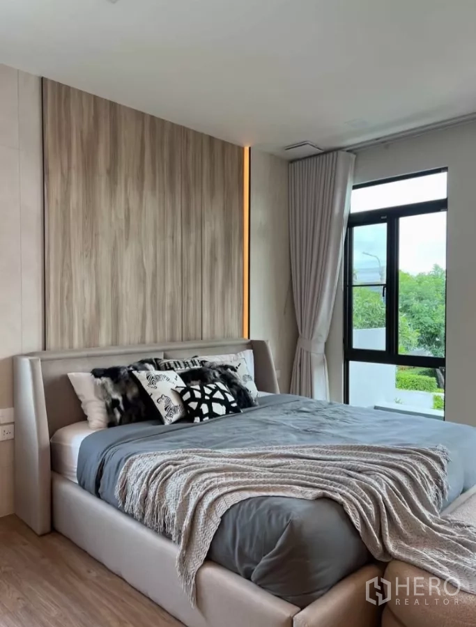 Detached House for rent in Samut Prakan - Master bedroom view with wood accent headboard, grey bedding and corner windows.