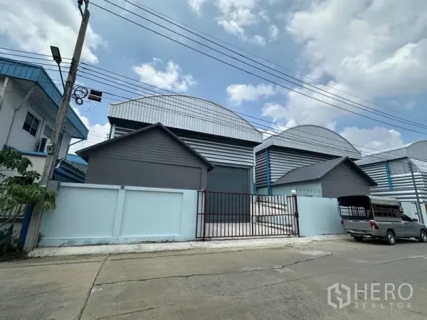 工厂 出租在Pak Kret, Nonthaburi - 工业区街景，小型工厂外观，拱形金属屋顶、浅蓝围墙与滑动大门