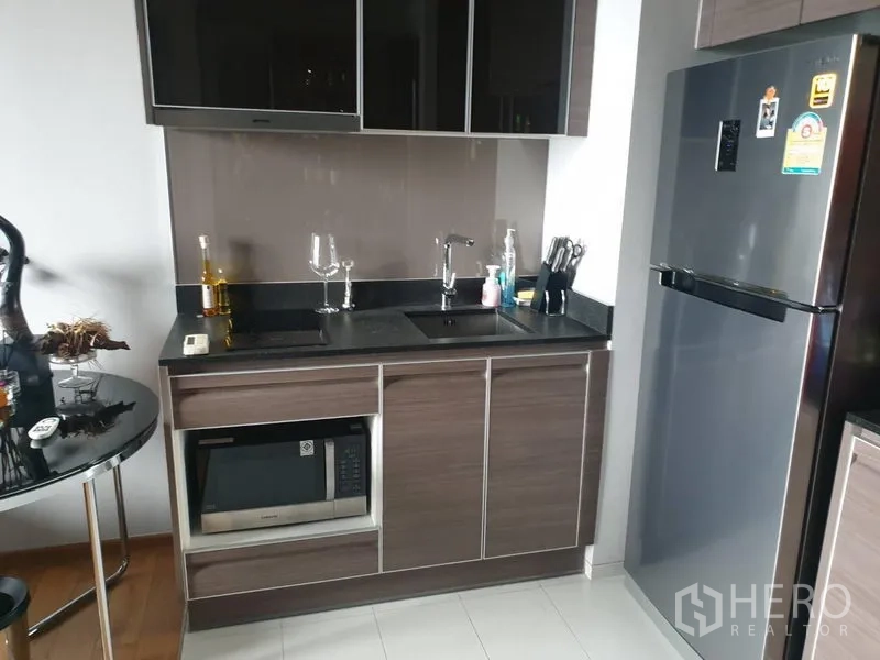 公寓 出租在Khlong Toei, Bangkok - 现代小厨房，亮面橱柜、黑色台面、水槽、微波炉与不锈钢冰箱。