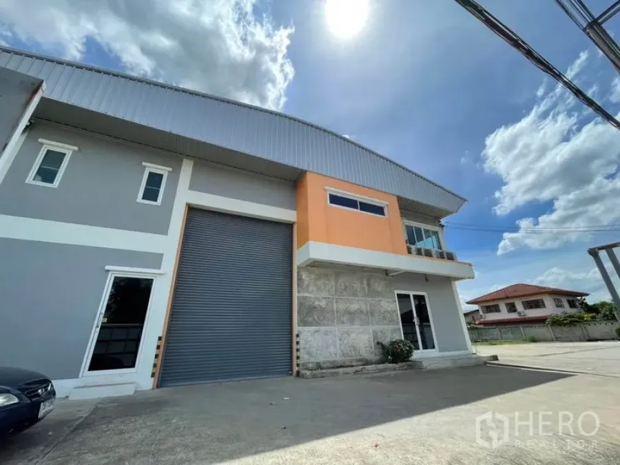 Warehouse for rent in Bang Kapi, Bangkok - Modern two-storey warehouse with grey roller shutter, orange facade and wide driveway under a sunny sky.