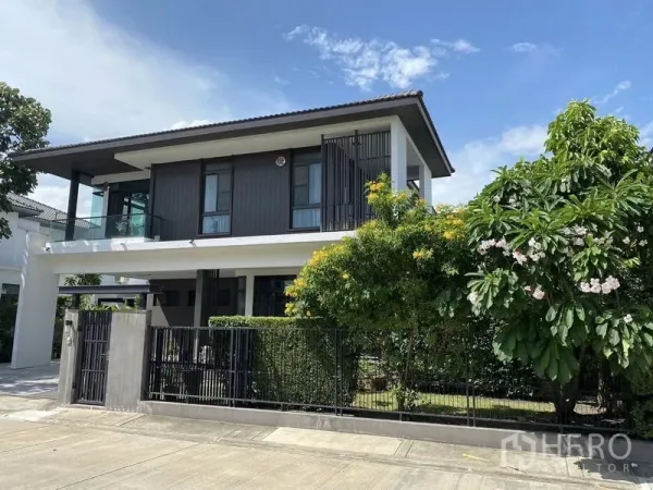 Detached House for rent or sale in Bang Phli, Samut Prakan - Street view of contemporary two-storey house behind black fence and lush greenery.