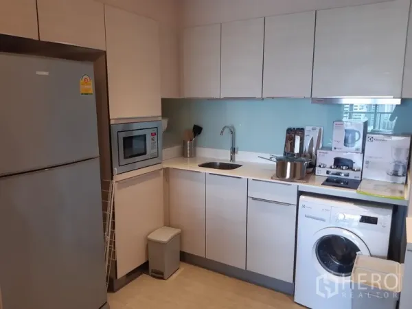 Condominium for rent in Watthana, Bangkok - Compact L-shaped kitchen with white cabinetry, fridge, microwave, glass backsplash and in-unit washing machine.