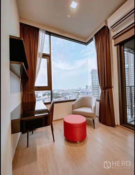 Condominium for sale in Huai Khwang, Bangkok - Corner work nook with desk, armchair, red ottoman and panoramic city view by the balcony door.